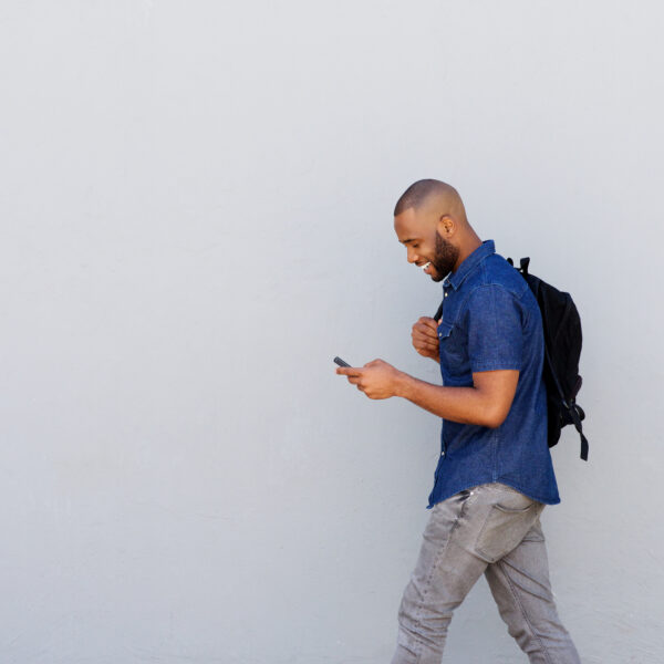 Side portrait of a happy young guy with bag walking outdoors and sending text message by mobile phone